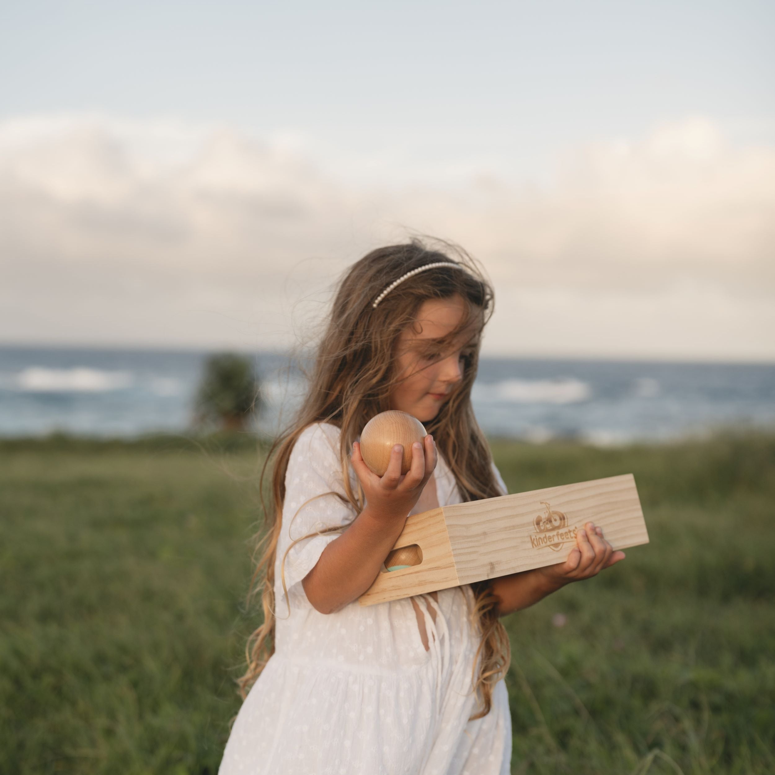 Young girl in a white dress holding a wooden box and a boule in a grassy field with ocean view.
